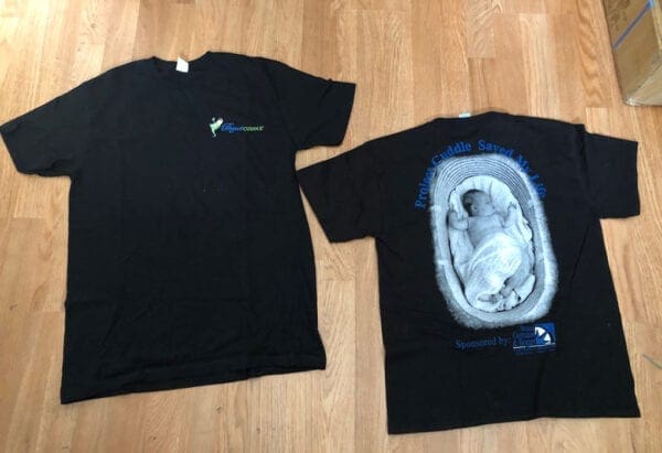Two Project Cuddle black t-shirts. Left (facing front): Project Cuddle logo; Right (facing back): Baby on a basket